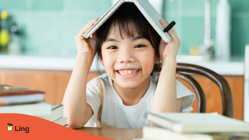 Thaimädchen sitzt am Schreibtisch und legt ein Buch über ihren Kopf beim lernen von Präpositionen auf Thai